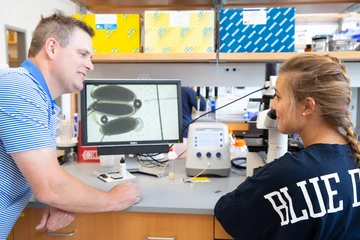a fredonia professor addreses a student as she examines material under a microscope