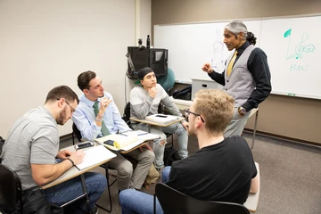 a professor engages with three students at their desk in a classroom