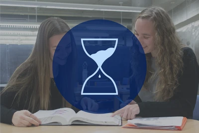 Two students work on project together in the classroom with an icon of an hourglass.