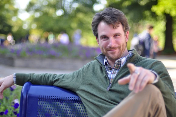 Ted Sharon sitting on a park bench smiling, hand on knee pointing at camera