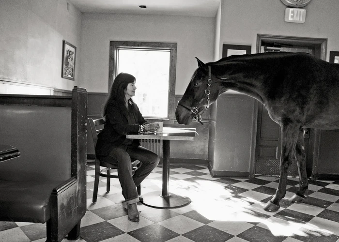 Black and white photograph of poet Joy Harjo sitting in a café facing a live horse. This is