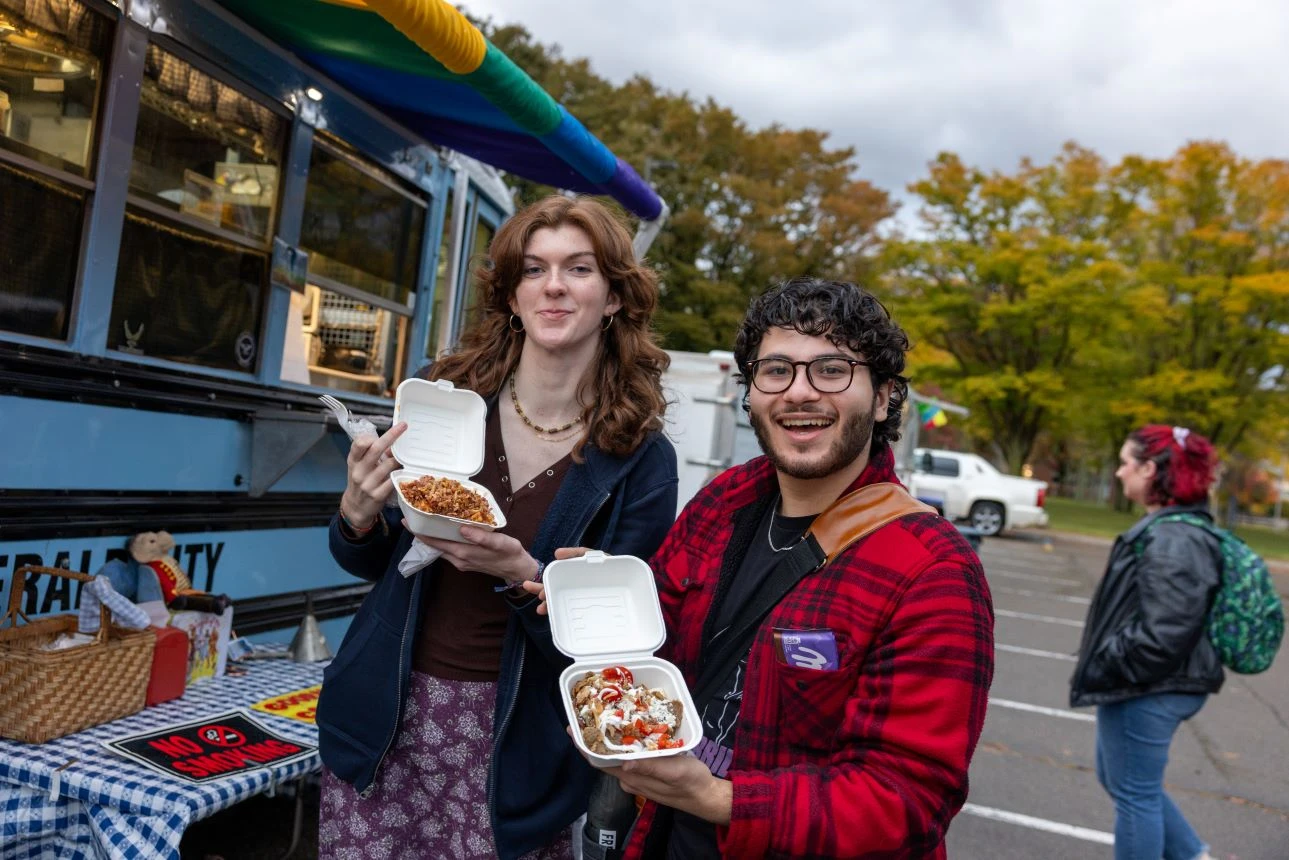 Two people display food containers in front of a food truck