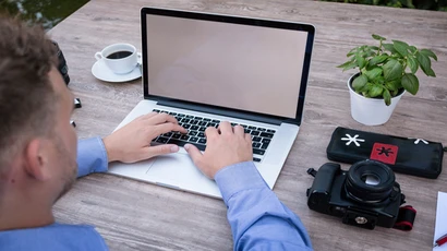 A man types on his laptop with a camera sitting next to the laptop. Degree in journalism, journalism degree, journalism major, journalism broadcasting.