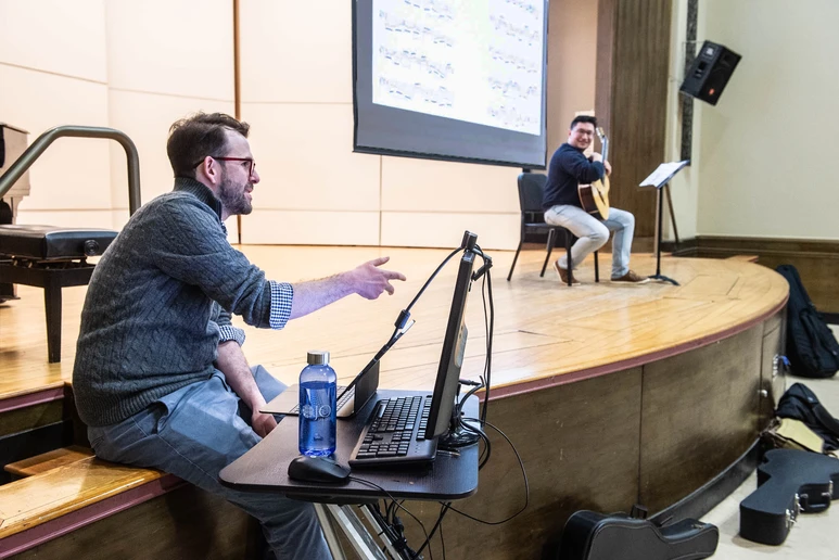 Dr. Nathan Huvard follows the score as Luke Villavicencio, a senior Music Performance major from Franklin Square, NY, plays on the Diers Recital Hall stage. The score is also projected onto an overhead screen for other students in the Guitar Recital Seminar to follow.